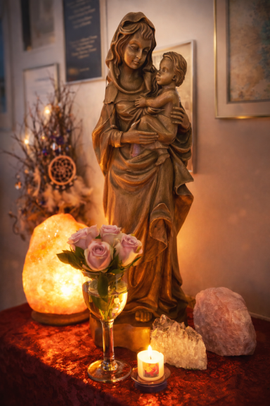 Statue der Jungfrau Maria mit Kind, umgeben von Kerzen und Blumen, auf einem dekorativen Tisch.