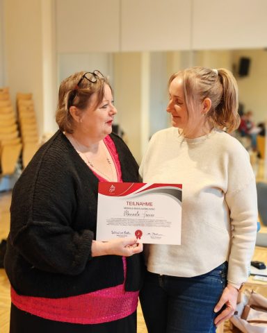 Zwei Frauenstehen sich gegenüber und halten ein Zertifikat in den Händen.